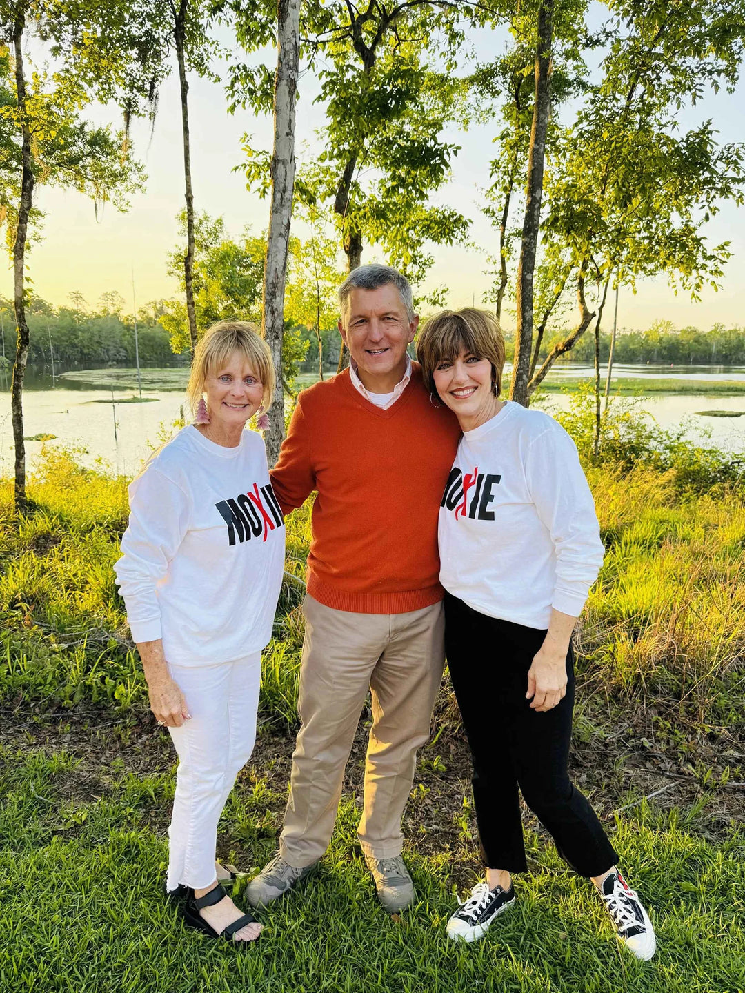Three people standing outdoors in a natural setting with trees and water in the background.