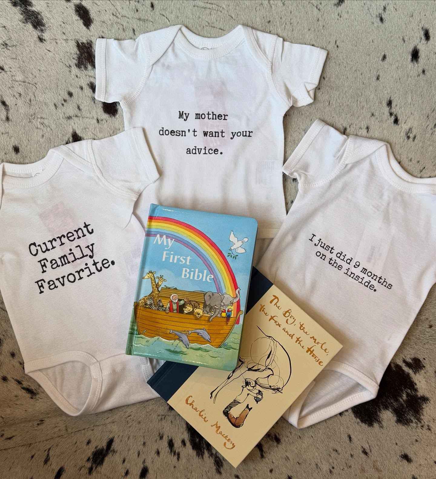 Three baby onesies with humorous text and a children's book on a textured surface.