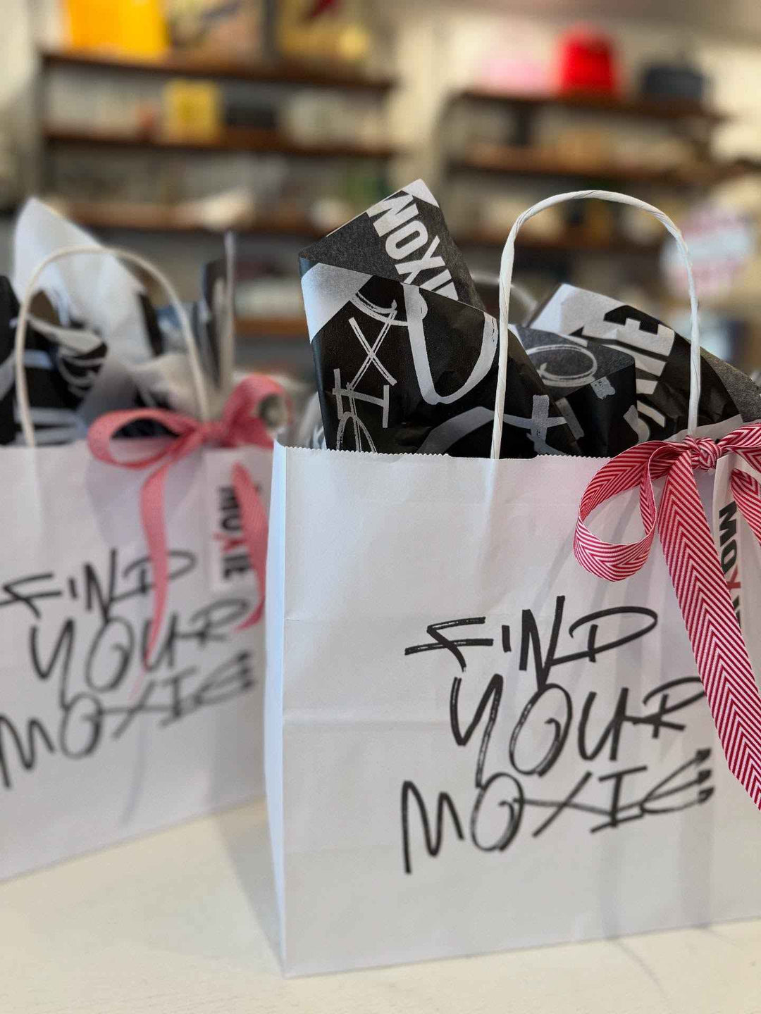White gift bag with 'Find Your Moxy' text, filled with black and white items, on a blurred background.