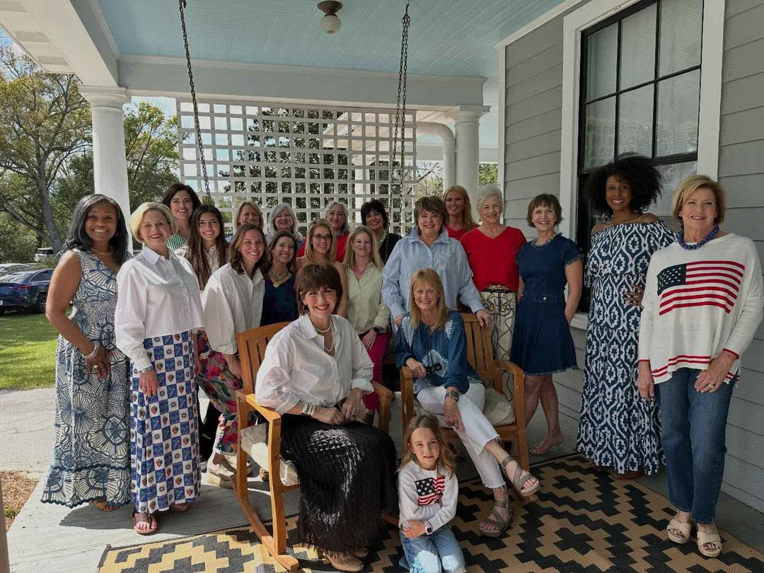 Group of people, including women and children, standing on a porch with a house and trees in the background.