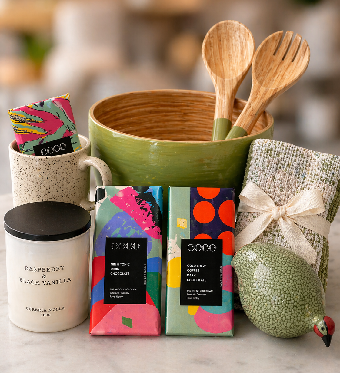 Kitchen items including a green bowl with wooden utensils, colorful COCO chocolate bars, and a candle on a marble surface.