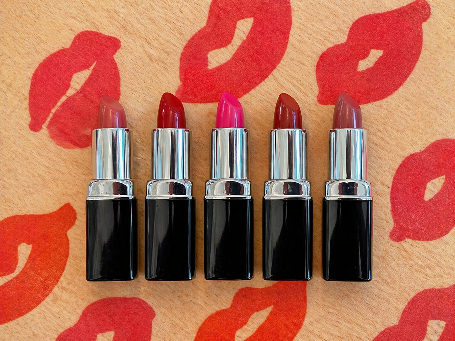 Five lipsticks with black cases on a beige background with red lipstick prints.