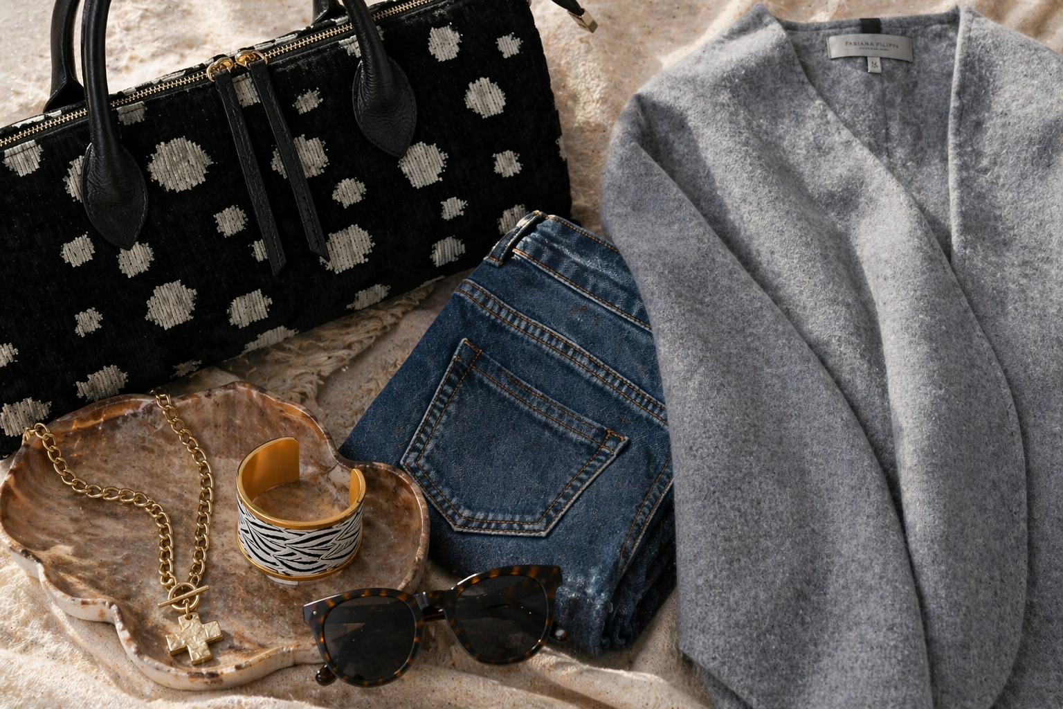 Black polka dot handbag, blue jeans, gray coat, sunglasses, and jewelry on a textured surface.