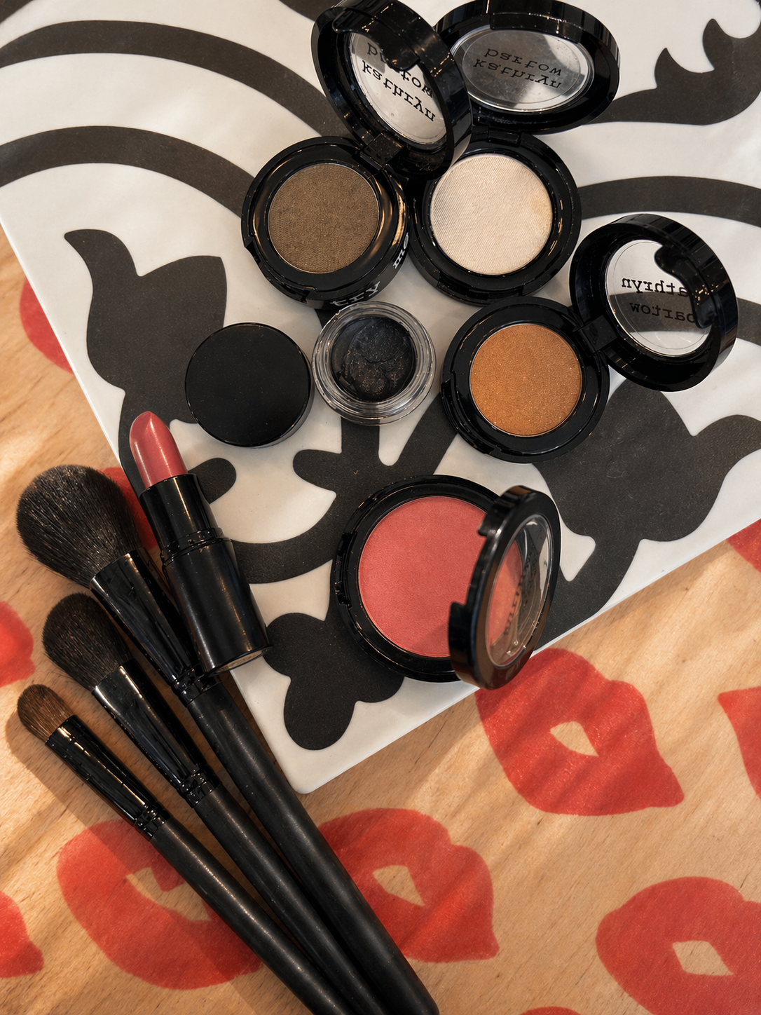 Makeup products including eyeshadows, a lipstick, and brushes on a patterned surface.
