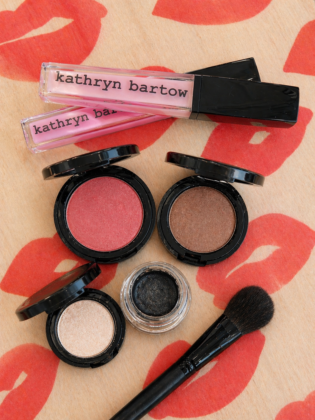 Makeup products including blush, eyeshadow, and lip gloss on a red and beige background.