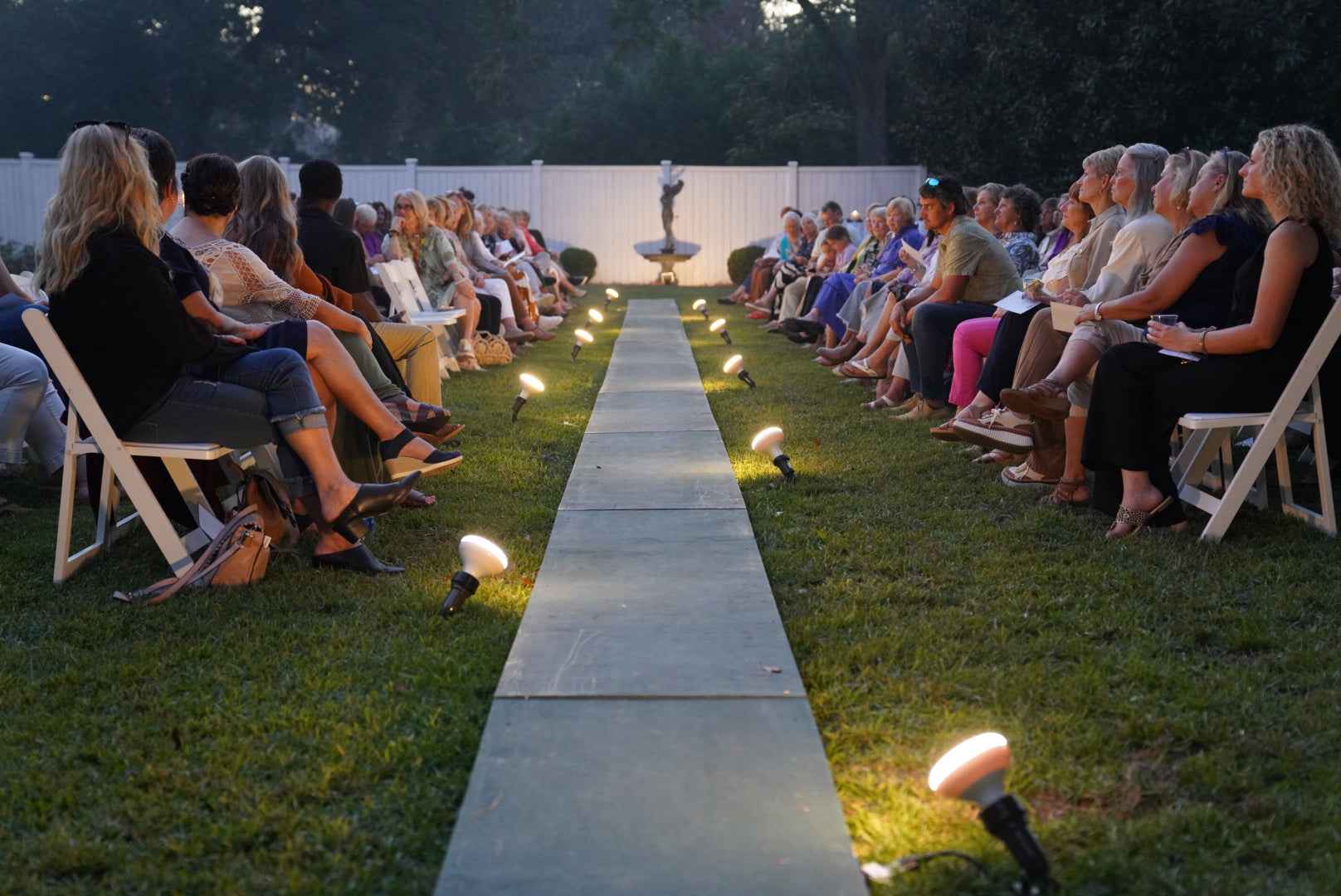 Audience seated on either side of a long, narrow pathway with decorative lights on a grassy area.