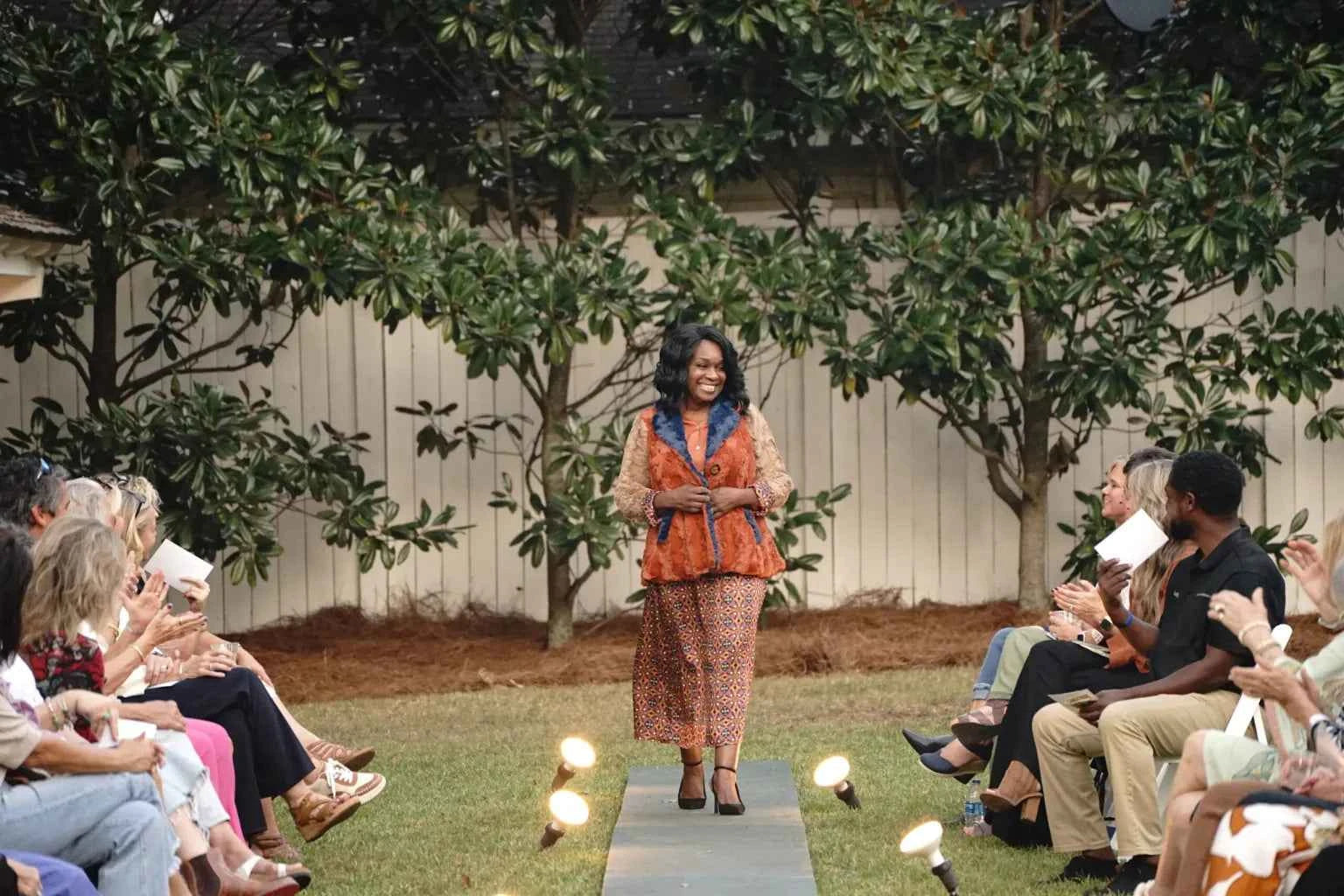 Woman in a patterned outfit standing on a grassy area with trees and people in the background.