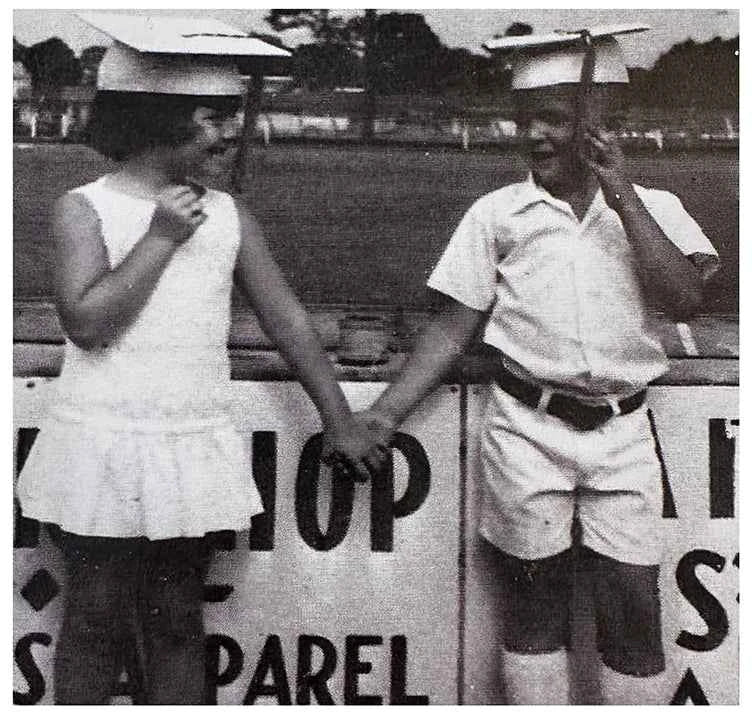 Two people in vintage attire standing next to a sign with text.